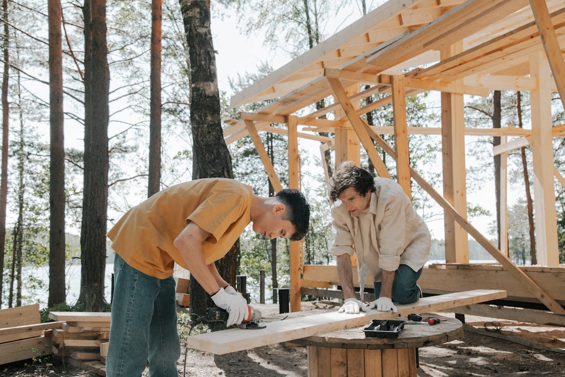 Two individuals working together to build a seasonal home, cutting wood and assembling materials outdoors in a forested setting