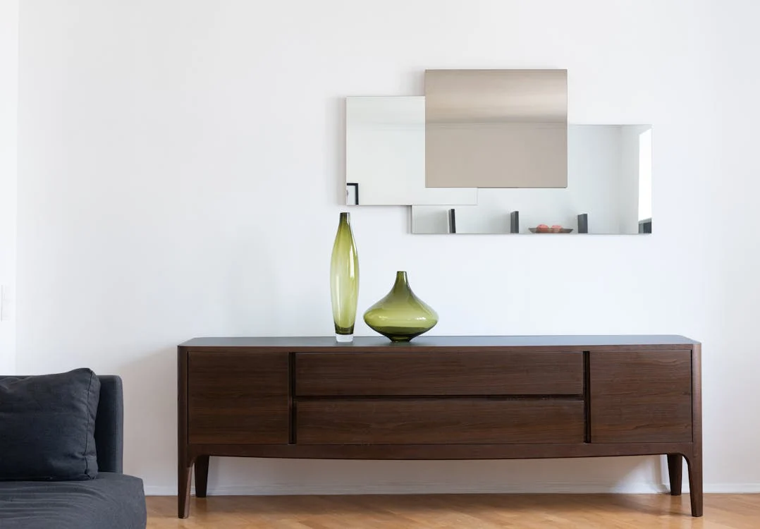 A cabinet with two green vases near a mirror on a white wall, illustrating a minimalist interior