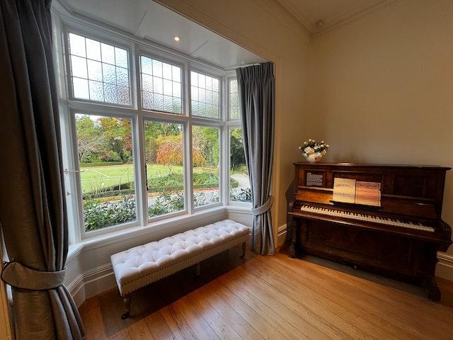 corner of a room with a piano and a bench standing in front of windows